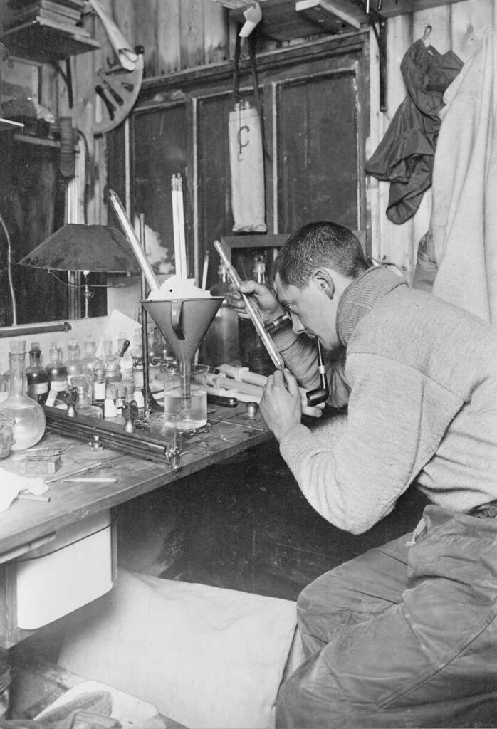 Edward W Nelson seated inside, at a laboratory table, testing thermometers during the British Antarctic ("Terra Nova") Expedition (1910-1913). Photograph taken on the 14th of July 1911, by Herbert George Ponting.
